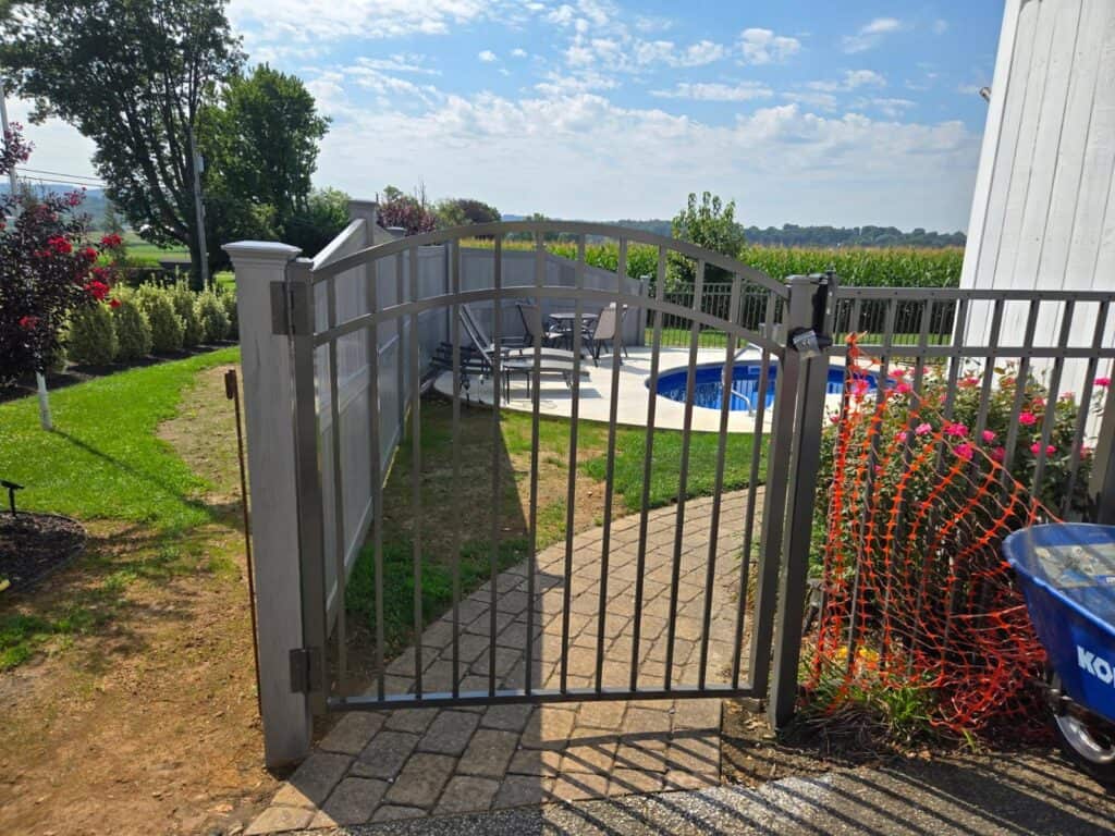Open black metal picket gate set between a grey solid privacy fence panel and a run of picket fencing, with a swimming pool and decking behind and a brick paver path through the gateway