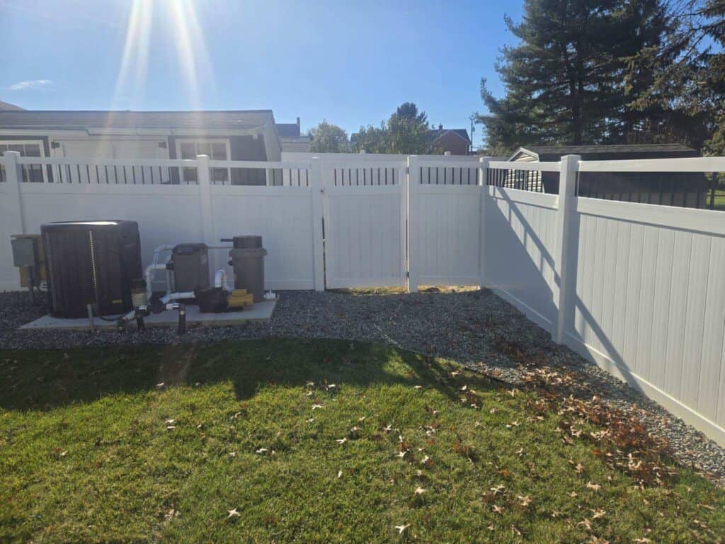 White vinyl privacy fence along the rear of a back yard with air conditioning units on a gravel pad, a gravel strip at the fence line and neighbouring houses beyond