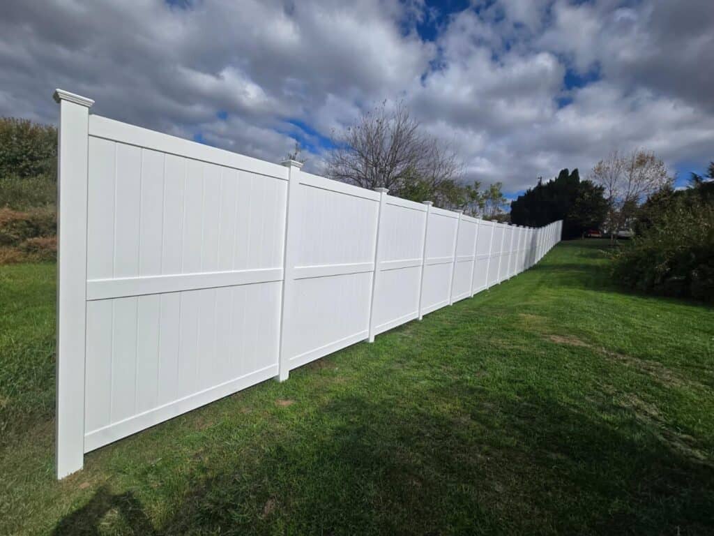 White vinyl privacy fence running the length of a mown lawn under a cloudy sky, with trees and shrubs along the boundary behind it